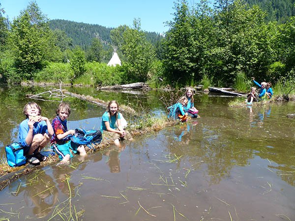 Twin Eagles Wilderness School summer campers wearing shorts with their legs in the murky waters of the shallow area of a lake, surrounded by trees with a view of hills in the far background.