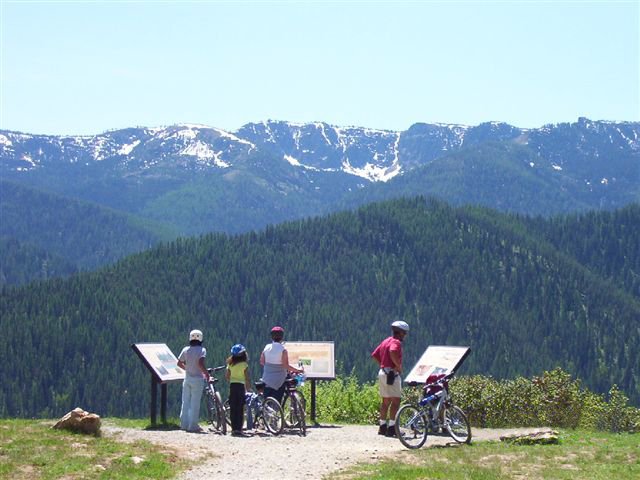 Bike riders taking a break to read historical marker signs along the Route of the Hiawatha, with a spectacular view of forest and mountain peaks in the distance.
