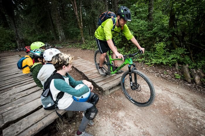 Man demonstrating mountain biking skills on a dirt trail to a group of students seated nearby on a wooden bench.