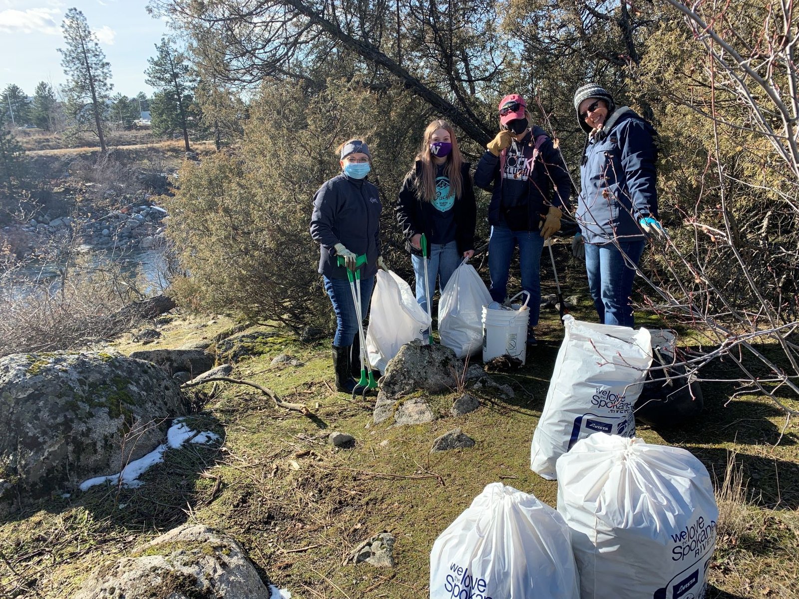 Four people volunteering during the Spokane River Clean-Up, wearing and holding pick-up pocking tools, and standing next to a large white bucket and four large plastic bags filled with collected trash.