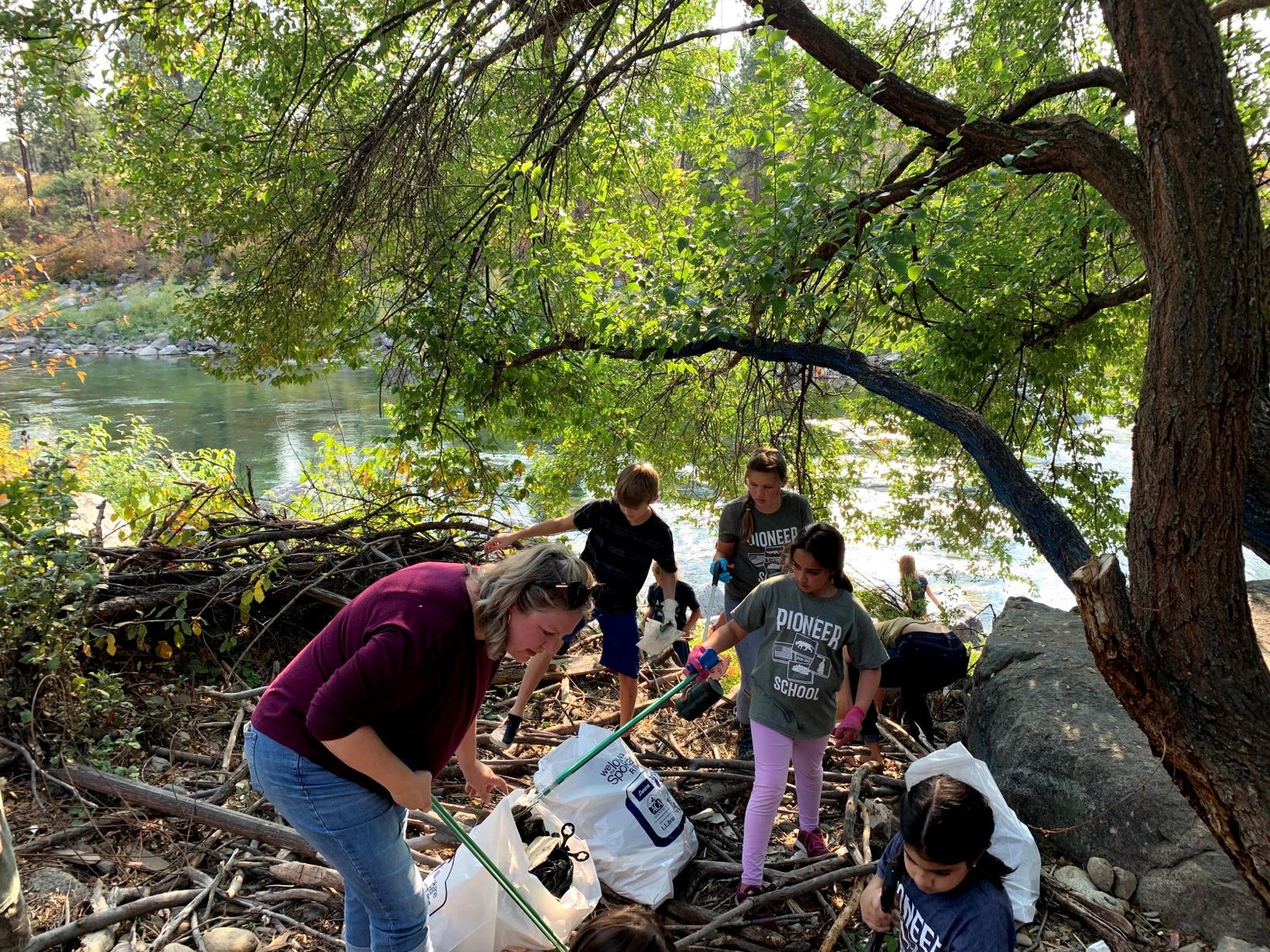 Volunteers, adults and youth wearing Pioneer School t-shirts, pick up garbage wearing gloves and using pick-up tools along the bank of the Spokane River.