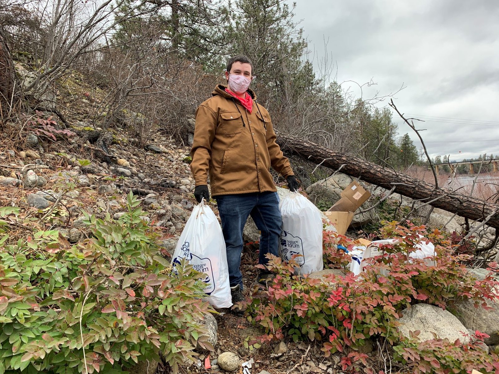 Man wearing a Carhart jacket, jeans, gloves, and facemask, carring two white plastic bags of litter and trash found along banks of the Spokane River.