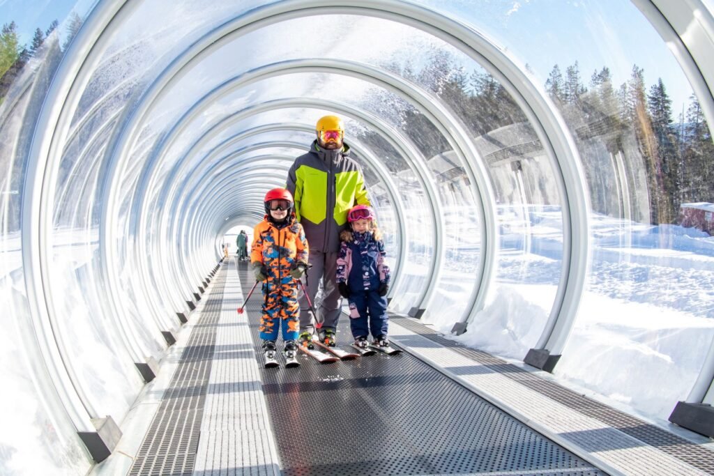 Adult and two children, all wearing ski gear, and standing on the Magic Carpet surface lift, enclosed in a clear tube.