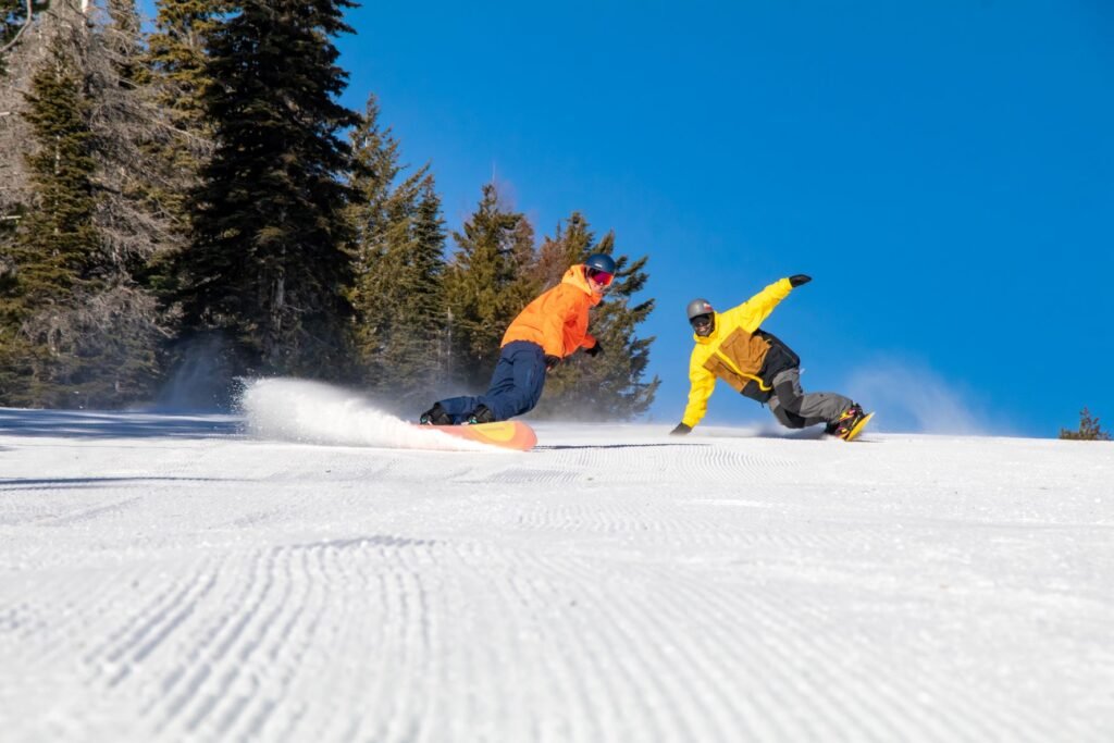 Two snowboarders, one wearing an orange jacket and the other in yellow, making big turns down a groomed run at 49 Degrees North.