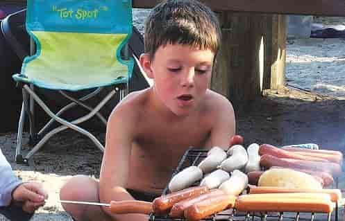 Young boy looking at the campfire grill covered in hotdogs and brats coving over the fire.