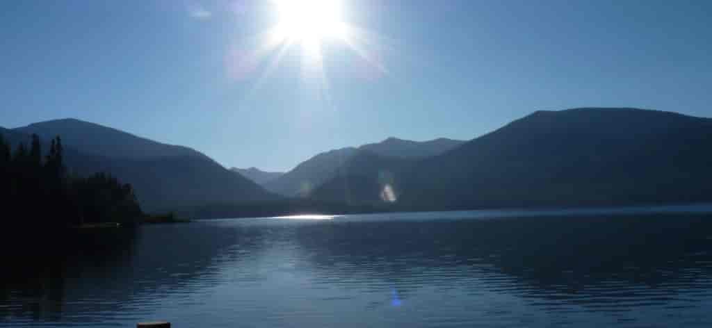 Still water of Priest Lake with mountain peaks and sun in the background.
